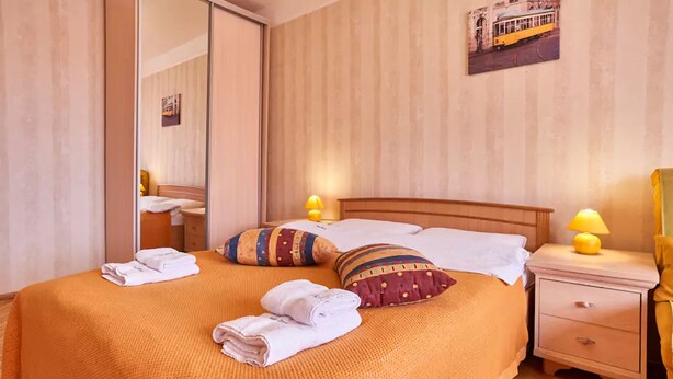 Ein ordentlich bezogenes Bett mit Holz-Kopfstück, zwei Kissen und zwei gefaltete Handtücher. Ein Nachttisch mit Lampe und Schublade ist vorhanden.