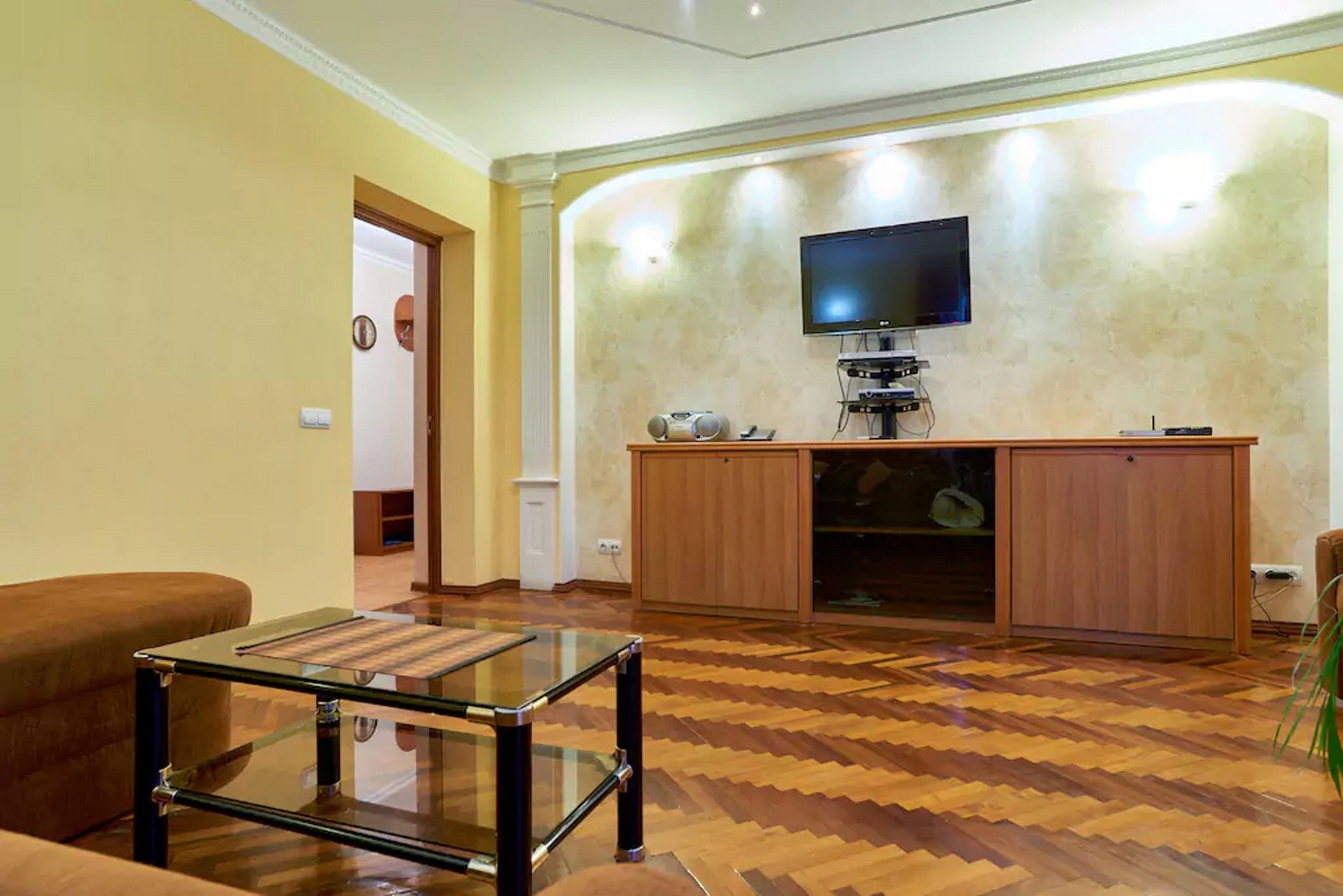 A living room with a wooden herringbone floor, a glass coffee table, a brown sofa, a wooden entertainment unit with a flat-screen TV, and a cabinet.