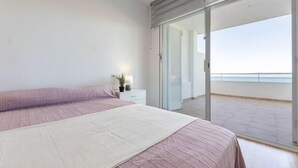 A modern bedroom with a large bed, a bedside table with a lamp, and a sliding glass door leading to a balcony with a view of the sea. - Apartment Rits by Interhome (Salou)