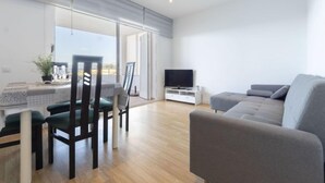 A modern living room with a grey sofa, a dining table with chairs, a TV on a white stand, and a large window with a view. - Apartment Rits by Interhome (Salou)
