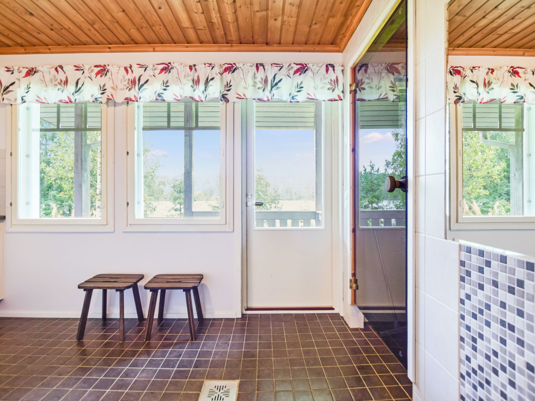 A covered patio area with a wooden ceiling, two windows with shutters, a white door, and a tiled floor.