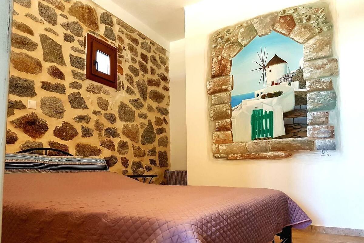A room with a stone wall, a bed with a quilt, a framed picture of a windmill, and a window with a view of the sea.