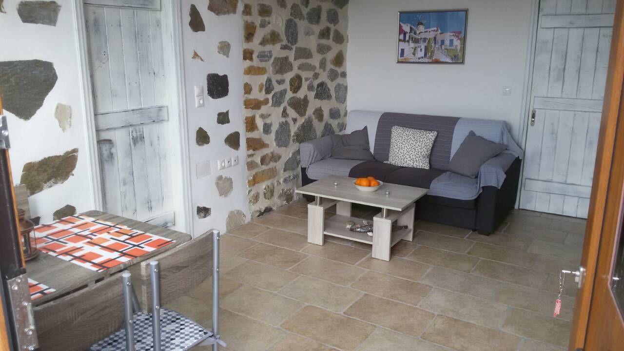 A cozy living room with a stone wall, a grey sofa, a small table with a bowl of fruit, and a checkered tablecloth.