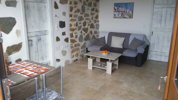 A cozy living room with a stone wall, a grey sofa, a small table with a bowl of fruit, and a checkered tablecloth.