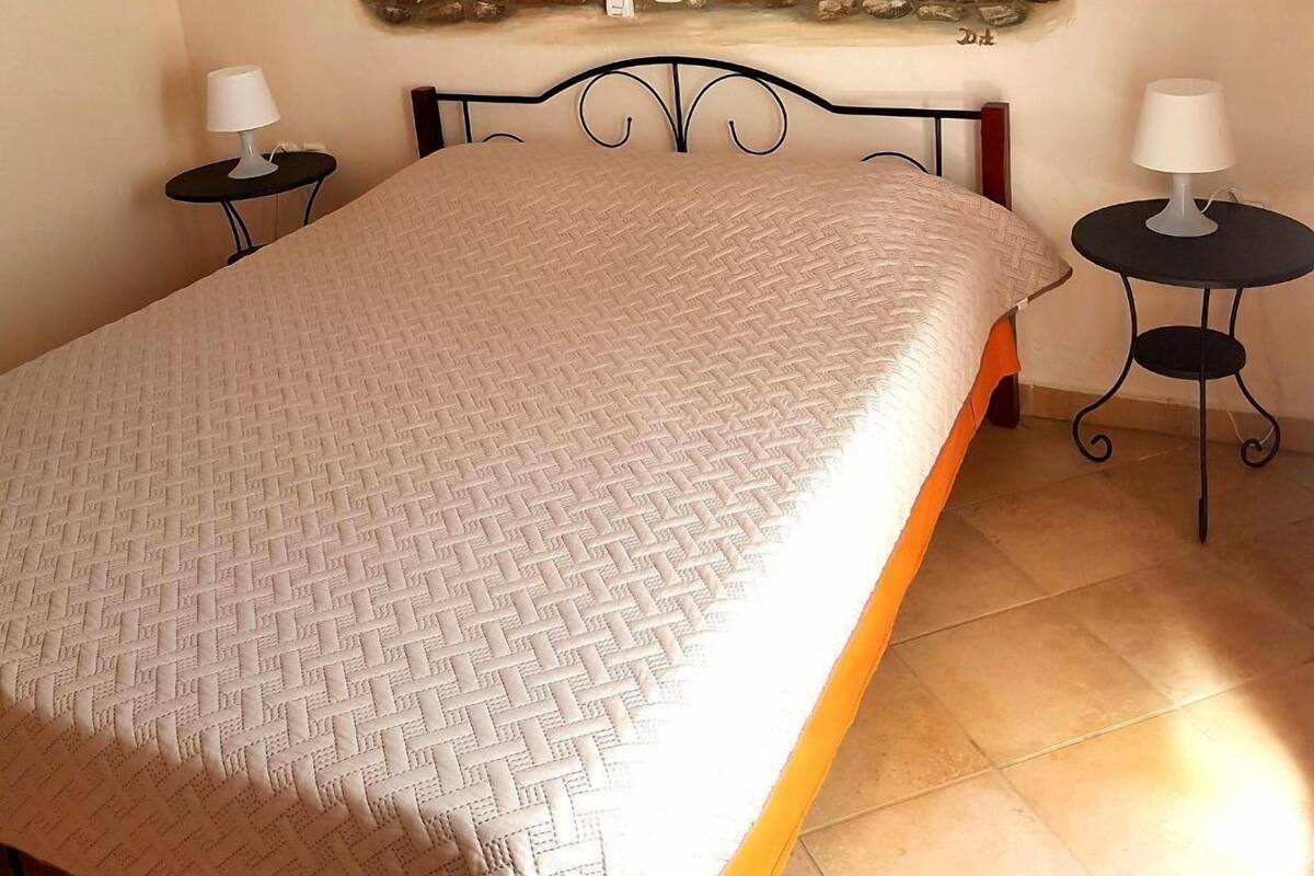 A bedroom with a bed, two bedside tables, a painting of a coastal scene, and a tiled floor.