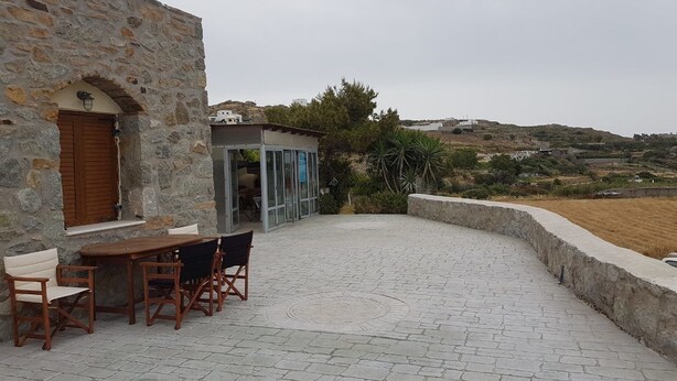 A stone building with a patio area featuring a wooden table and chairs, a glass enclosure, and a view of the countryside. - The Stone House Naxos (Naxos)