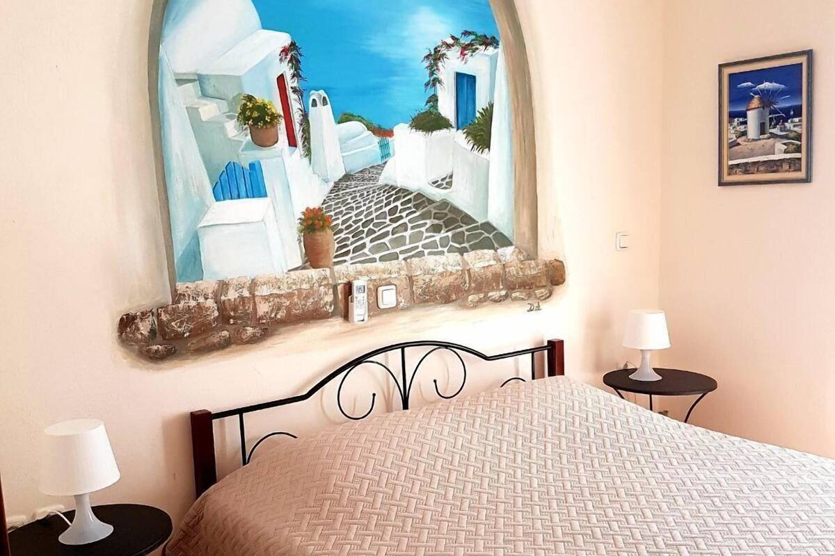 A bedroom with a bed, two bedside tables, a wall-mounted painting of a coastal scene, and a framed picture on the wall.
