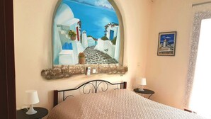 A bedroom with a bed, two bedside tables, a painting of a coastal scene, and a window with a curtain. - The Stone House Naxos (Naxos)