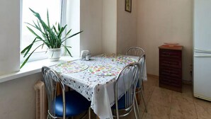 A dining area with a table covered by a patterned tablecloth, surrounded by metal chairs. A potted plant is on the windowsill, and a clock is on the wall. - Home-Hotel Streletskaya 28 (Kyiv)