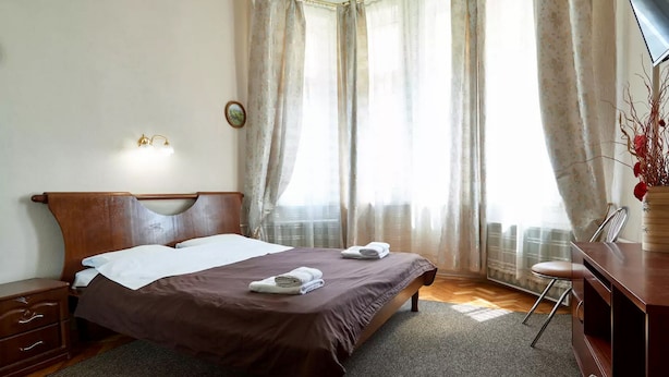 A neatly made bed with a brown bedspread and white pillows, a wooden nightstand with drawers, a desk with a chair, and a vase with red flowers on the right. - Home-Hotel Streletskaya 28 (Kyiv)