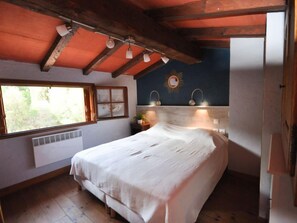 A bedroom with a wooden ceiling, a bed with a white bedspread, a window with a view of greenery, and a wall-mounted light fixture.