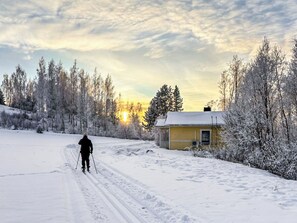 Una persona practica esquí de fondo en un sendero cubierto de nieve, con una casa y árboles al fondo.