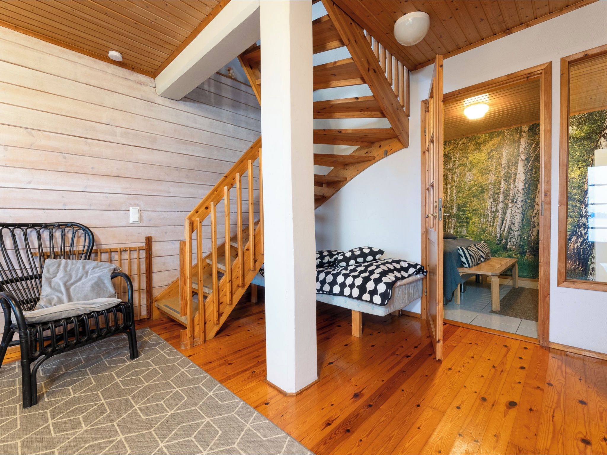 Una scala in legno conduce a una camera da letto soppalcata con un letto e vista su una foresta attraverso una porta vetrata.