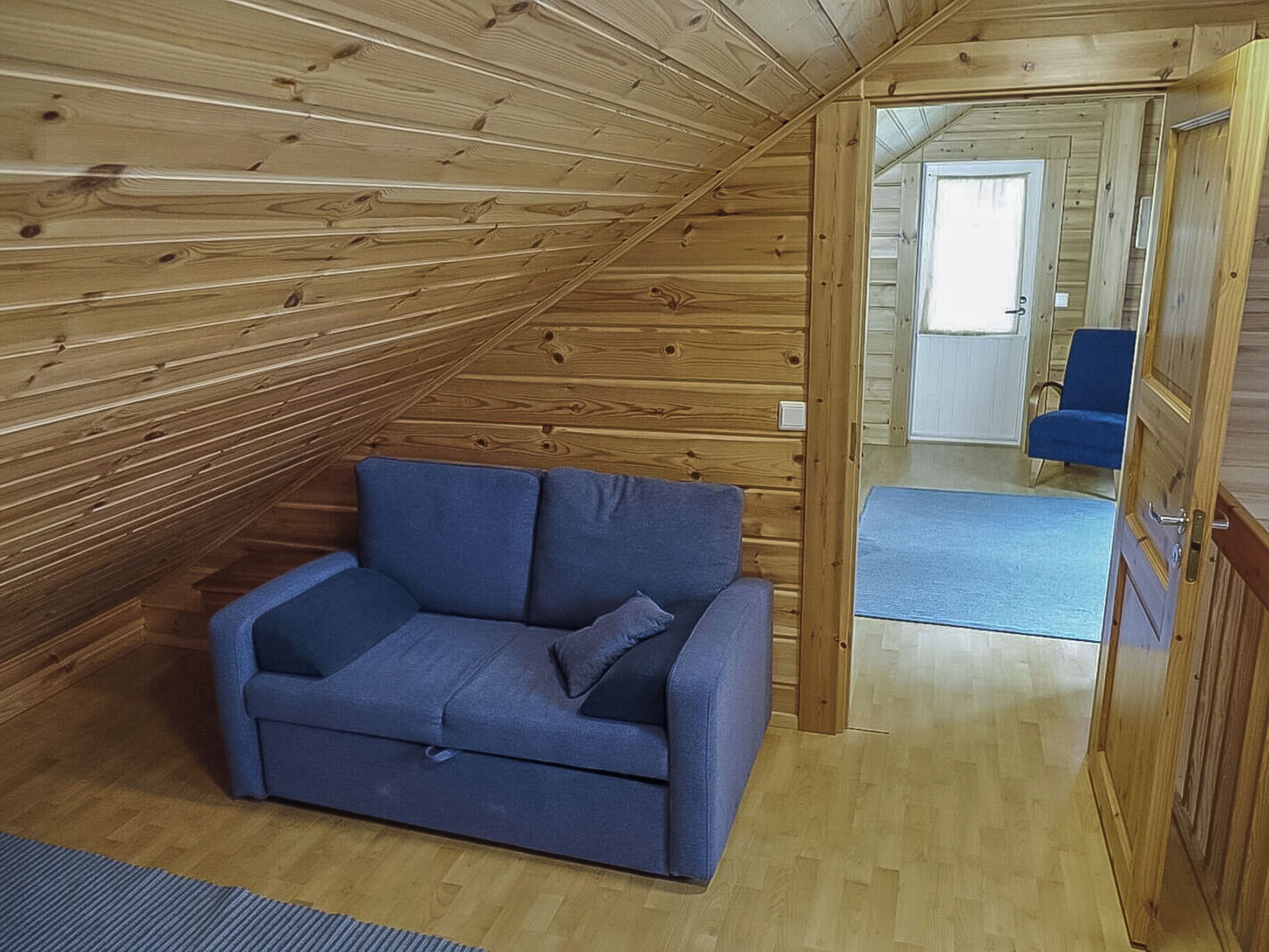 Huoneessa on puinen, kalteva katto, sininen sohva ja ovi, joka johtaa toiseen huoneeseen.