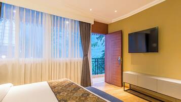 A hotel room with a large bed, a TV mounted on the wall, and a balcony with a view of greenery.