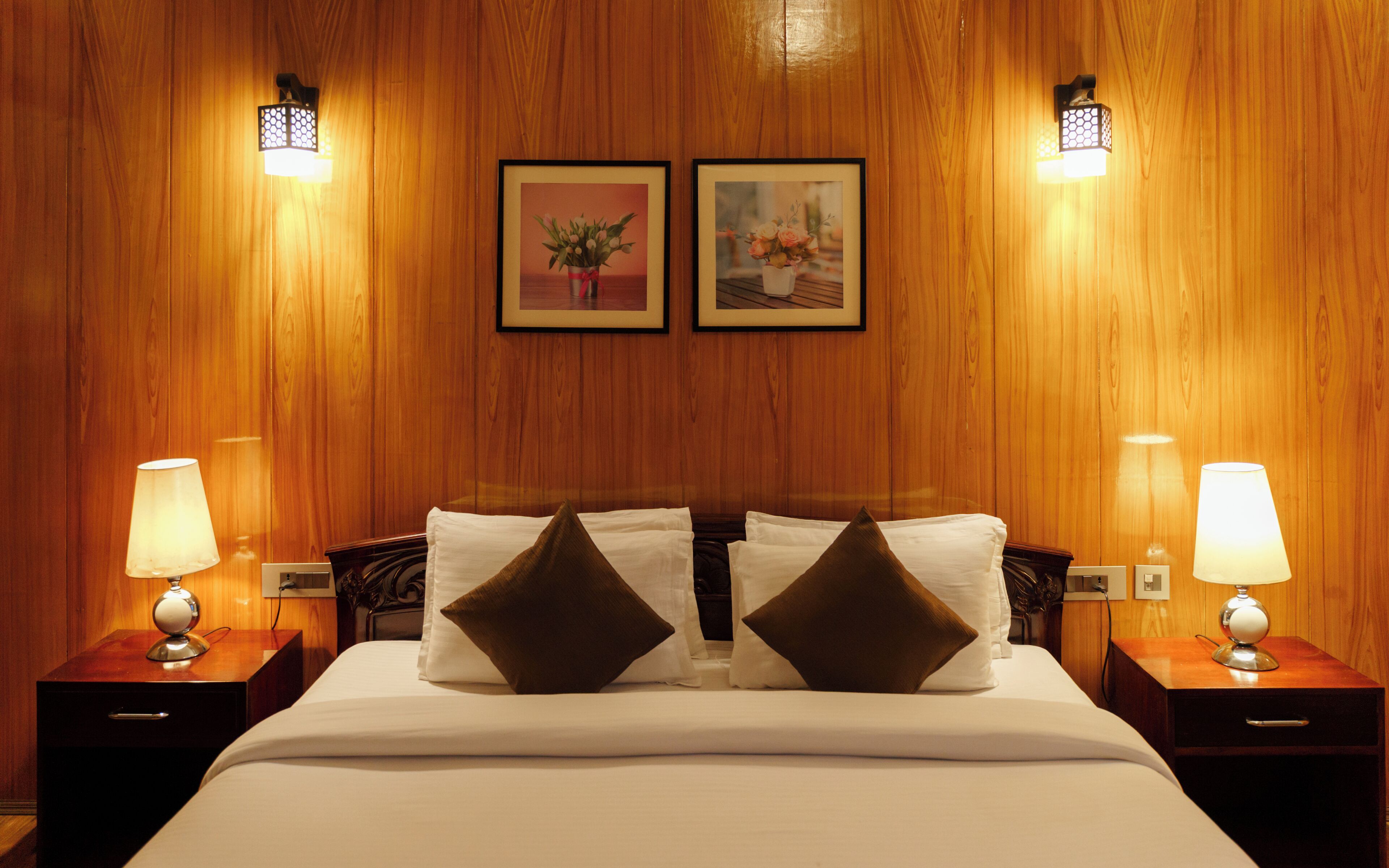 Un cuarto de hotel con pared de paneles de madera, una cama grande con ropa blanca y dos almohadas marrones, dos lámparas de noche y dos cuadros en la pared.