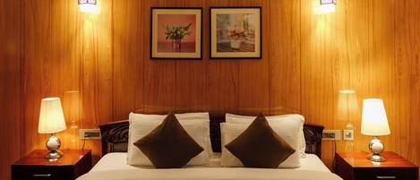Un cuarto de hotel con pared de paneles de madera, una cama grande con ropa blanca y dos almohadas marrones, dos lámparas de noche y dos cuadros en la pared.