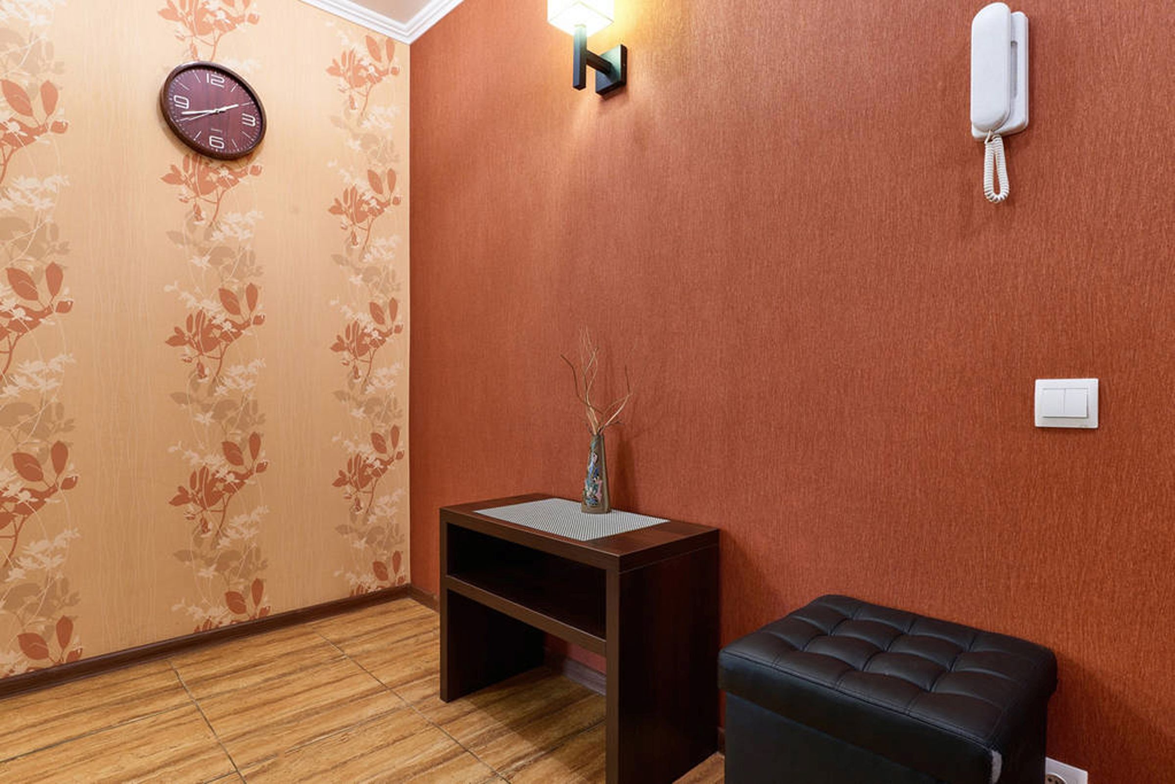 A room with a wooden floor, a small wooden table with a vase, a black leather ottoman, a wall clock, a wall-mounted light, and a phone.