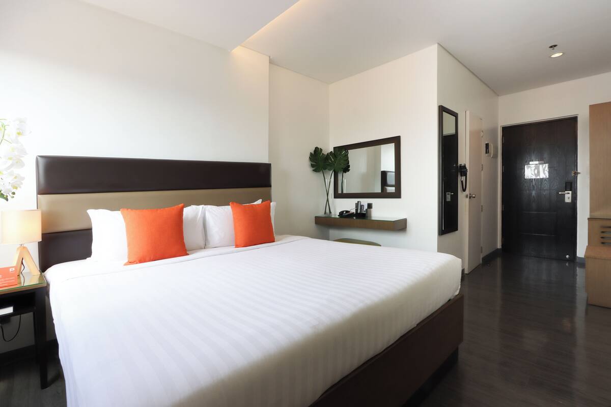 A modern hotel room with a large bed, a nightstand, a mirror, and a small desk with a lamp.
