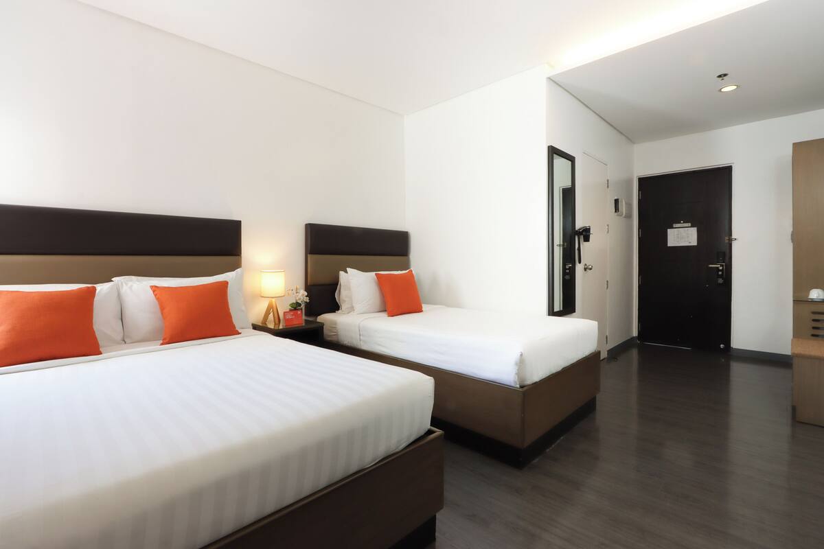 A hotel room with two beds, each with white bedding and orange pillows, a wooden headboard, a nightstand with a lamp, and a door leading to another room.