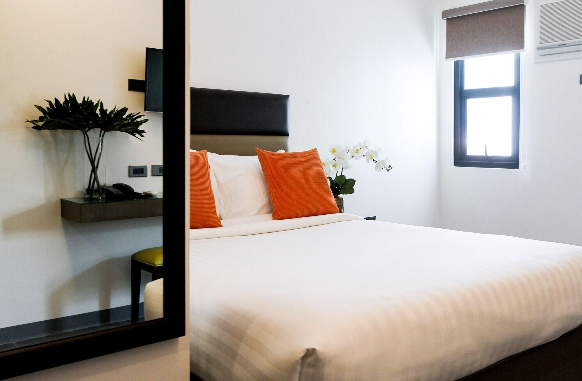 A hotel room with a large bed, a bedside table with a vase of flowers, a small stool, and a window with blinds.