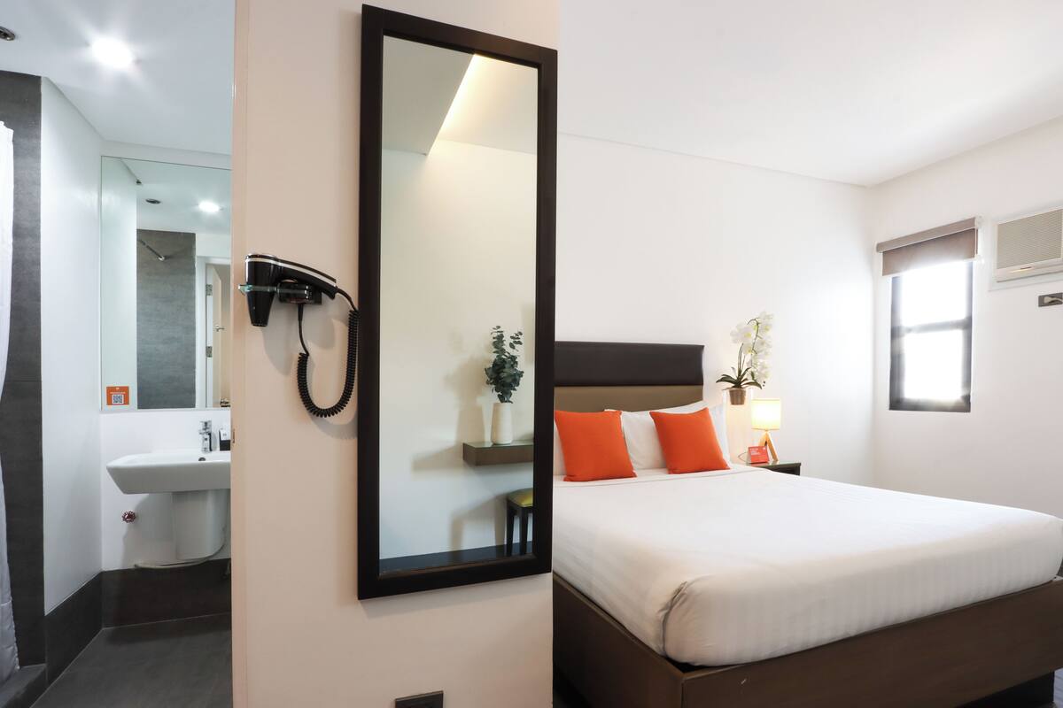 A hotel room with a large bed, a bedside table with a lamp, a mirror, a bathroom with a sink, and a shower area.