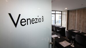 A conference room with tables and chairs, a white wall with the text "Venezia B" visible, and large windows allowing natural light. - Privato Makati (Makati)
