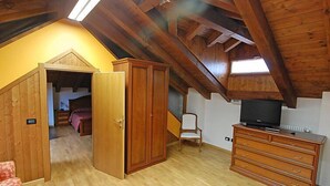 Quarto com teto inclinado, vigas de madeira, cama, guarda-roupa, televisão e cadeira.