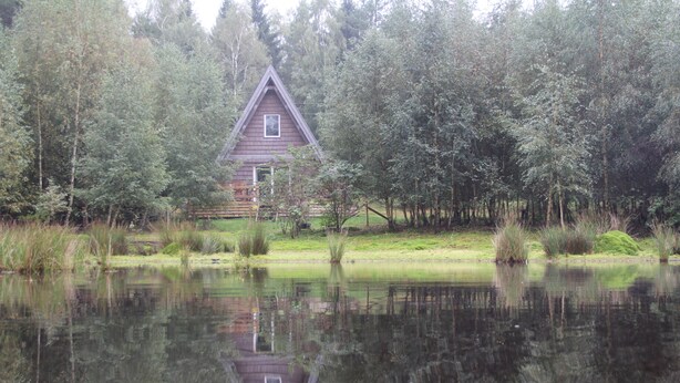 A cabin with a triangular roof, surrounded by trees and a body of water. - Äventyrscampen in Sövde (Sjobo)