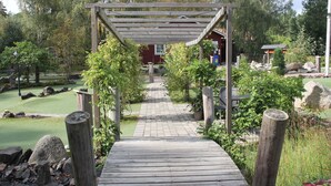 A wooden pathway leading to a gazebo in a landscaped garden with a red building in the background. - Äventyrscampen in Sövde (Sjobo)