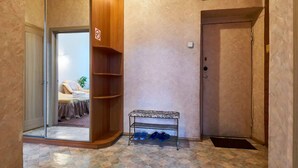 Un couloir avec un banc en bois, un porte-chaussures et une porte donnant sur une pièce où se trouvent un lit et une chaise.