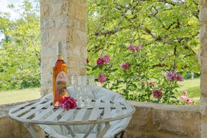 A table with a bottle and glasses, a white chair, and a garden with pink flowers. - Beautiful typical French Manor with private swimming pool (Anglars Nozac)