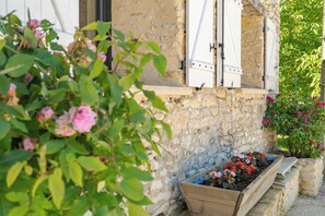 Un bâtiment en pierre avec des volets en bois, un parterre fleuri avec différentes espèces de fleurs et un banc en bois.