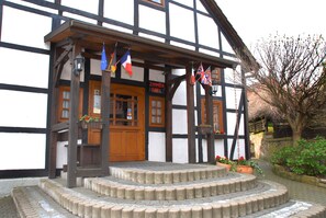 A traditional half-timbered building with a wooden door and steps leading to the entrance. Flags are hanging from the roof, and there are flower pots on the steps. - Hotel Alte Schule (Luhden)