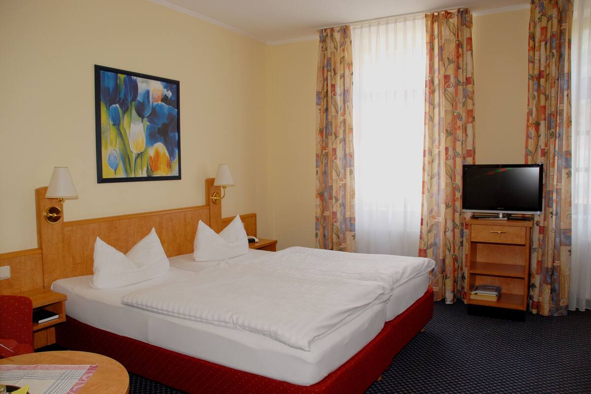 A hotel room with a large bed, two bedside tables with lamps, a television on a stand, a painting on the wall, and a window with curtains.