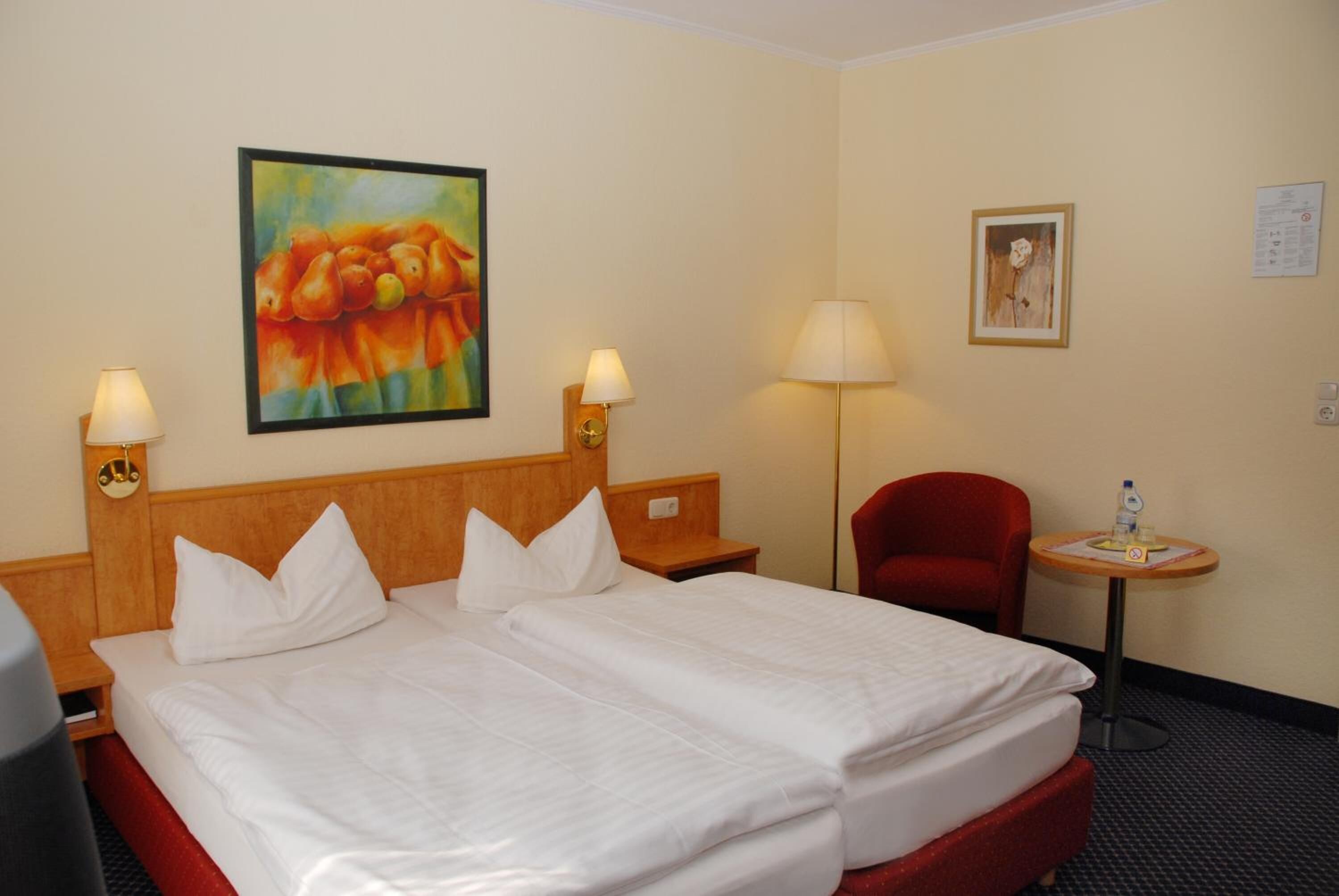 A hotel room with a double bed, two bedside tables, a red armchair, a small round table with a lamp, and two wall-mounted lamps.