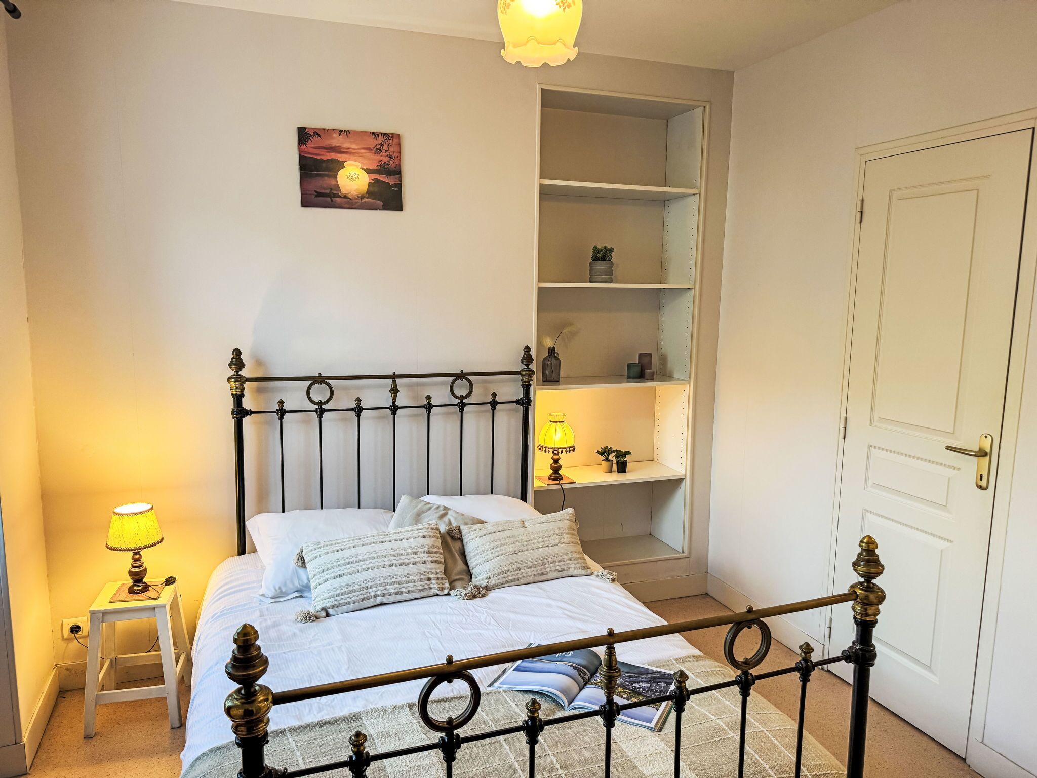 A bedroom with a bed, bedside lamps, a shelf unit, and a door.