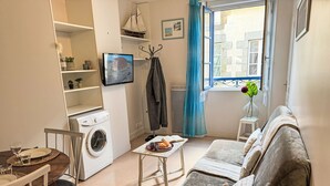 A compact living space with a sofa, a dining table with chairs, a washing machine, a small table with a book and a plant, and a window with curtains. - Le Vieux Port by Interhome (Cancale)