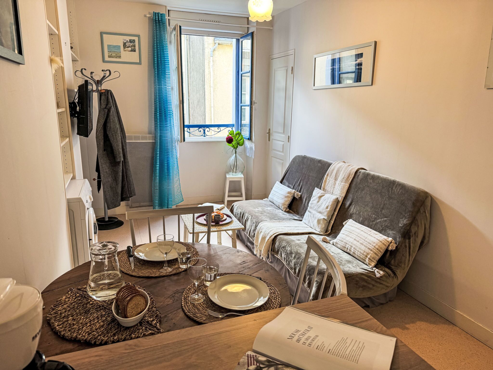Ein gemütliches Wohnzimmer mit einem grauen Sofa, einem kleinen Esstisch und einem Fenster mit Vorhängen.