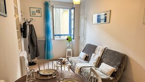 A cozy living room with a grey sofa, a small table set for dining, and a window with curtains. - Le Vieux Port by Interhome (Cancale)