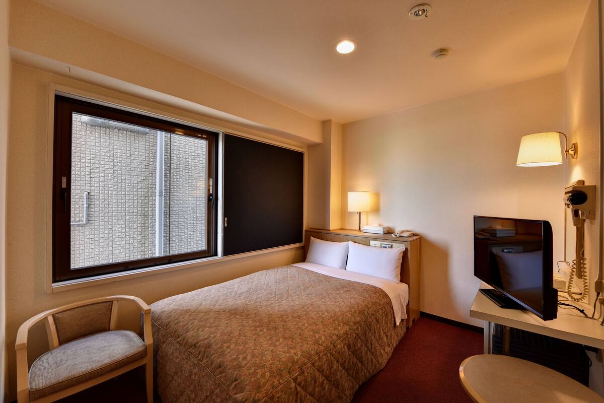Une chambre d’hôtel comprenant un lit, une chaise, un bureau avec une lampe, une télévision et une fenêtre avec des stores.