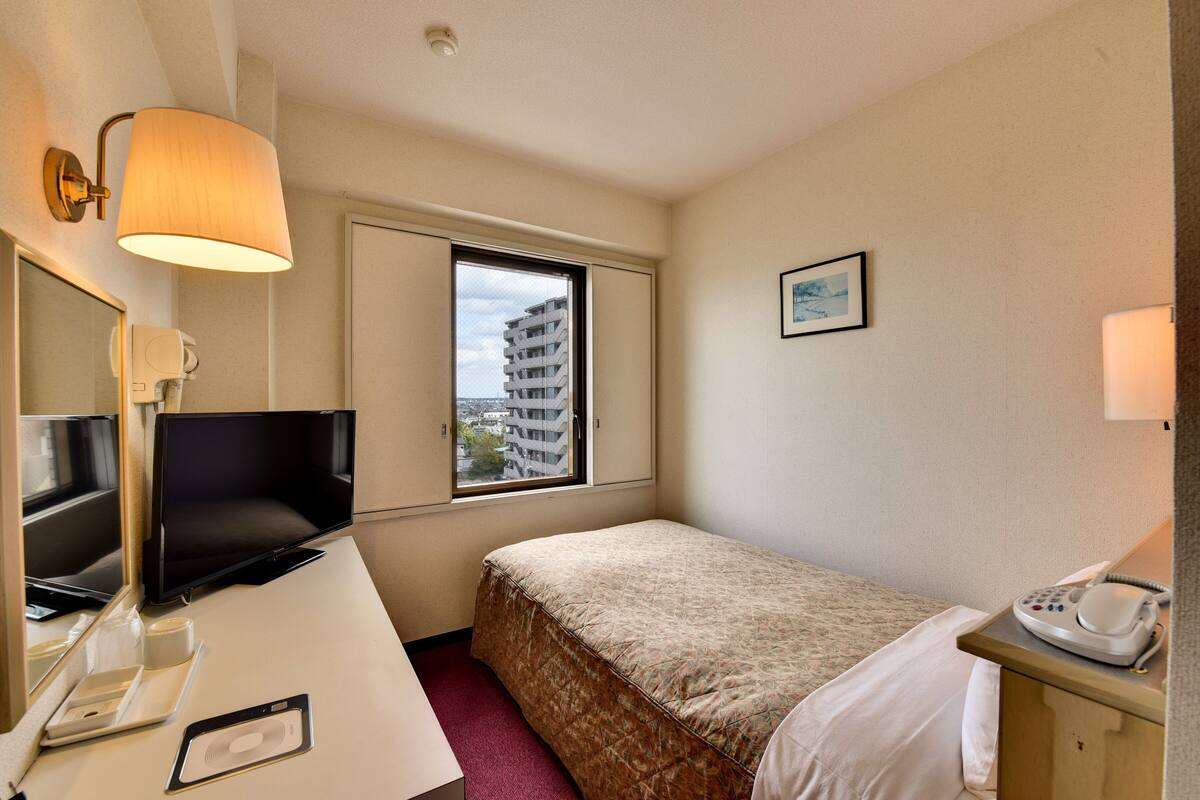 Une chambre d’hôtel comprenant un lit, un bureau avec un ordinateur, un téléphone, une fenêtre donnant sur des immeubles et une lampe fixée au mur.