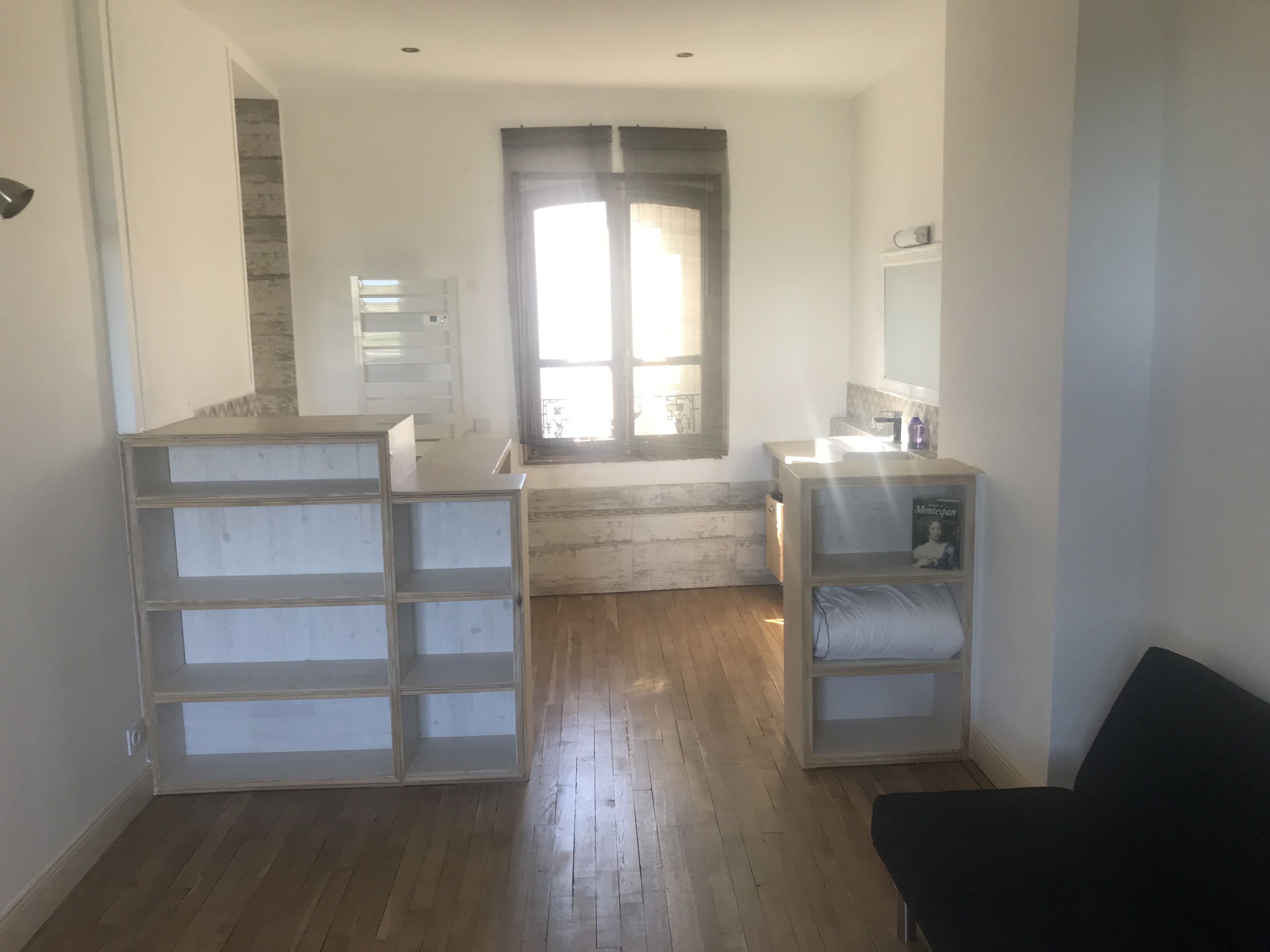A room with a wooden floor, a black chair, a white shelf unit, and a window with curtains.