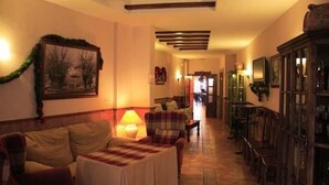 A cozy living room with a sofa, armchair, and a small table with a lamp. There is a fireplace and a bookshelf in the background. - Hotel Patri (Huescar)