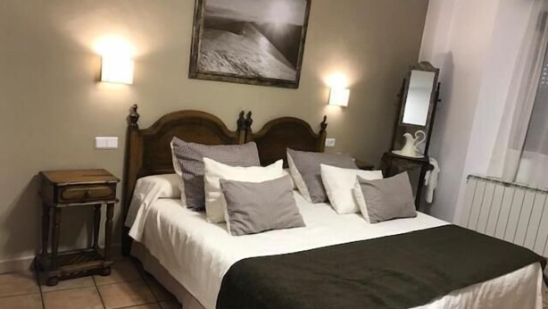 A neatly made bed with a wooden headboard, two bedside tables, a nightstand, a wall-mounted mirror, and a framed picture above the bed. - Hotel Patri (Huescar)