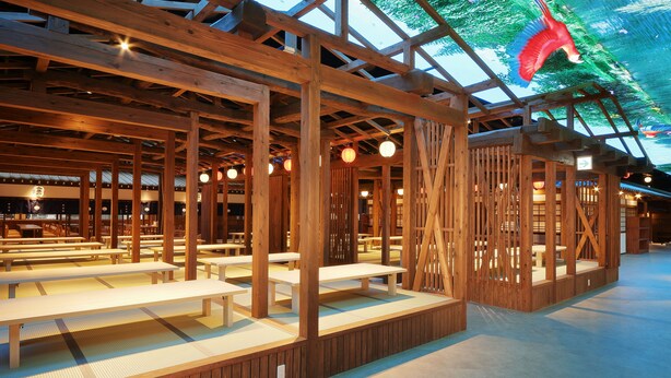 A spacious indoor area with wooden benches and tables, a high ceiling with wooden beams, and a large digital screen displaying colorful graphics. - Yurakujo (Tomisato)