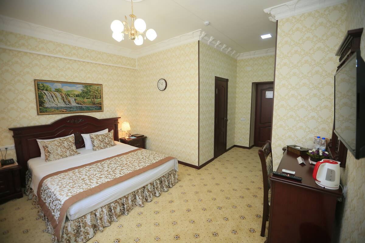 Een hotelkamer met een groot bed, een nachtkastje met een lamp, een klok aan de muur en een bureau met een waterkoker en afstandsbediening.