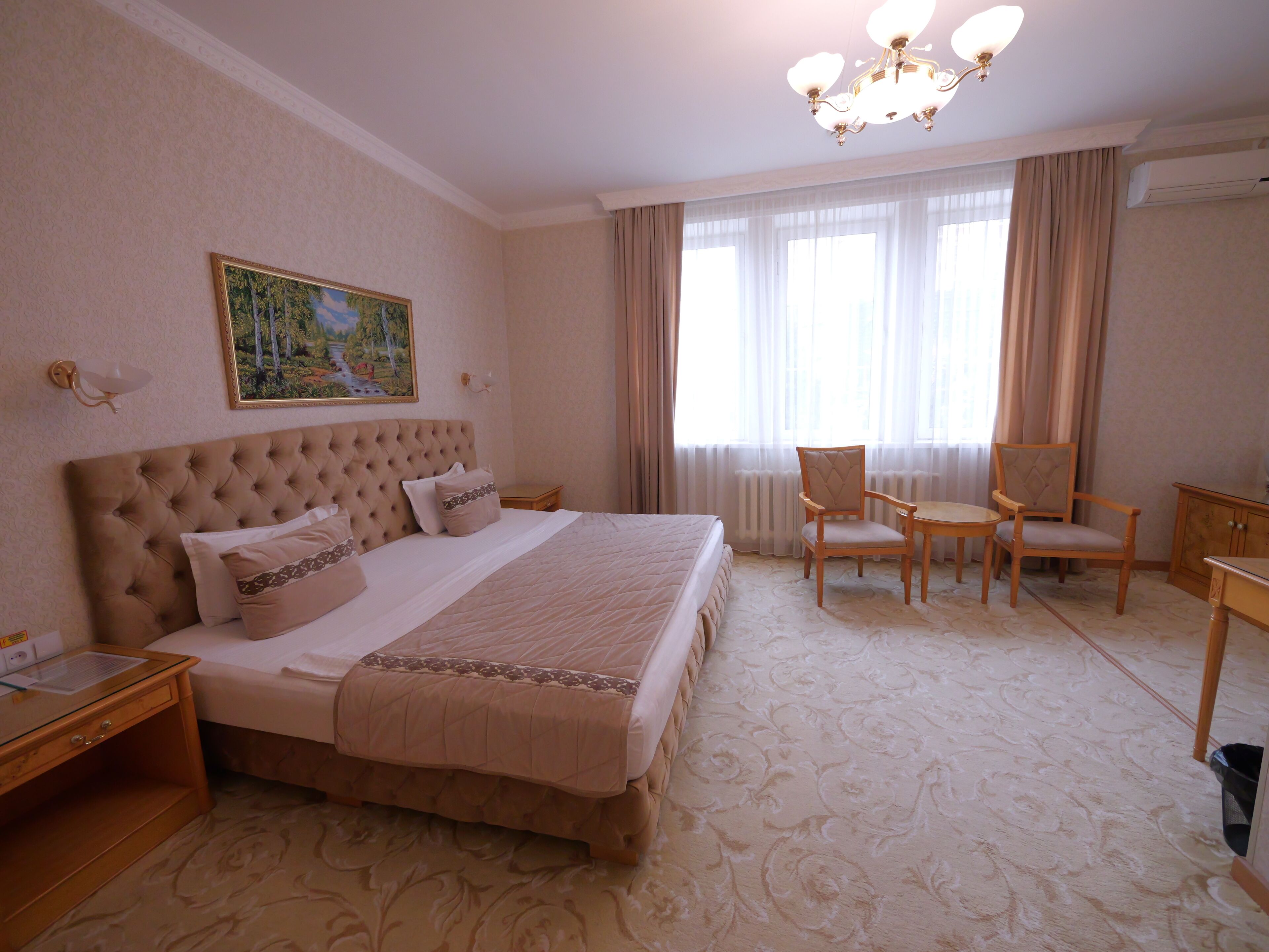 A hotel room with a large bed, a desk, two chairs, a chandelier, and a painting on the wall.