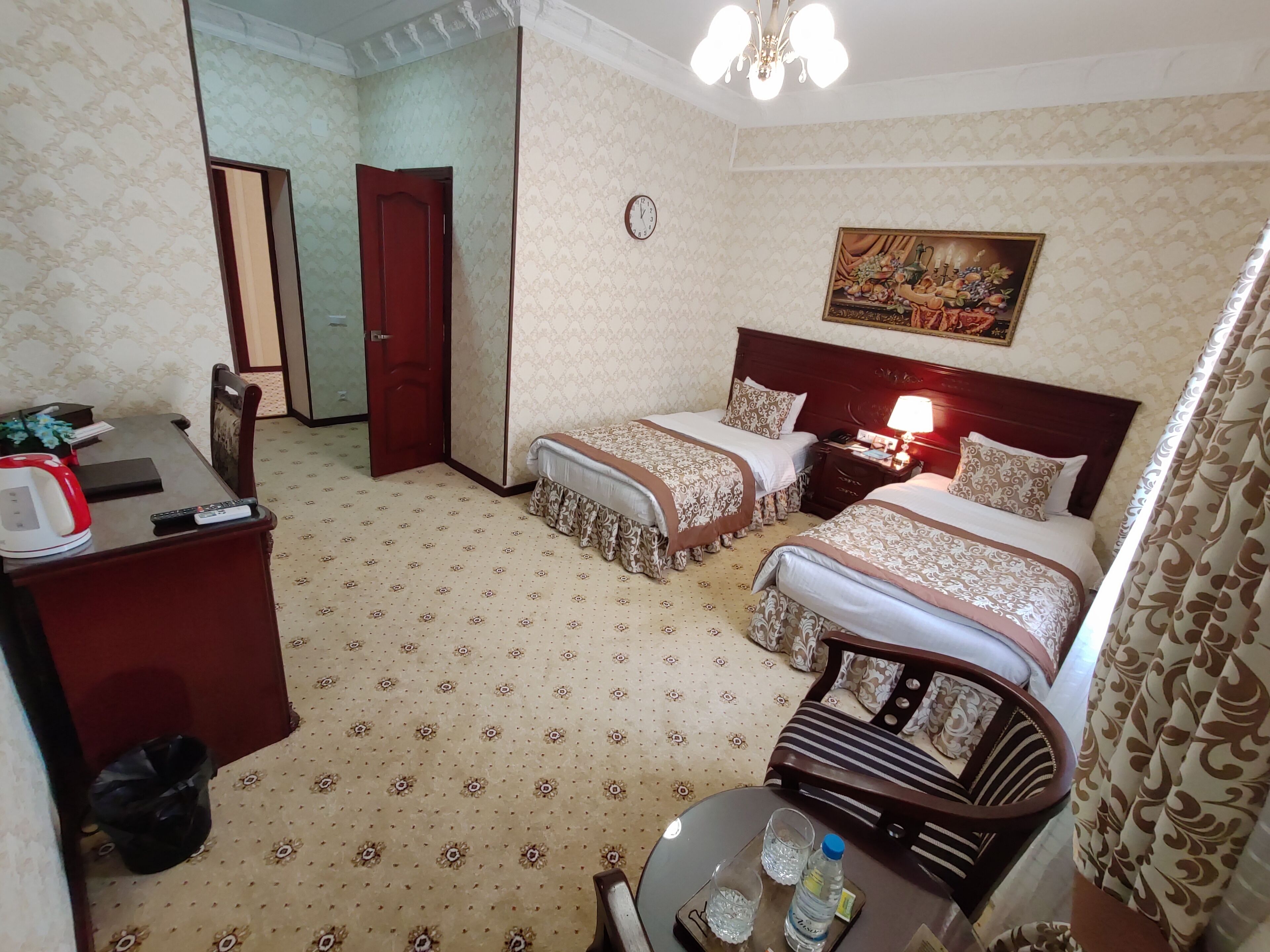 A hotel room with two beds, a desk with a kettle, a trash bin, and a table with glasses and a bottle.