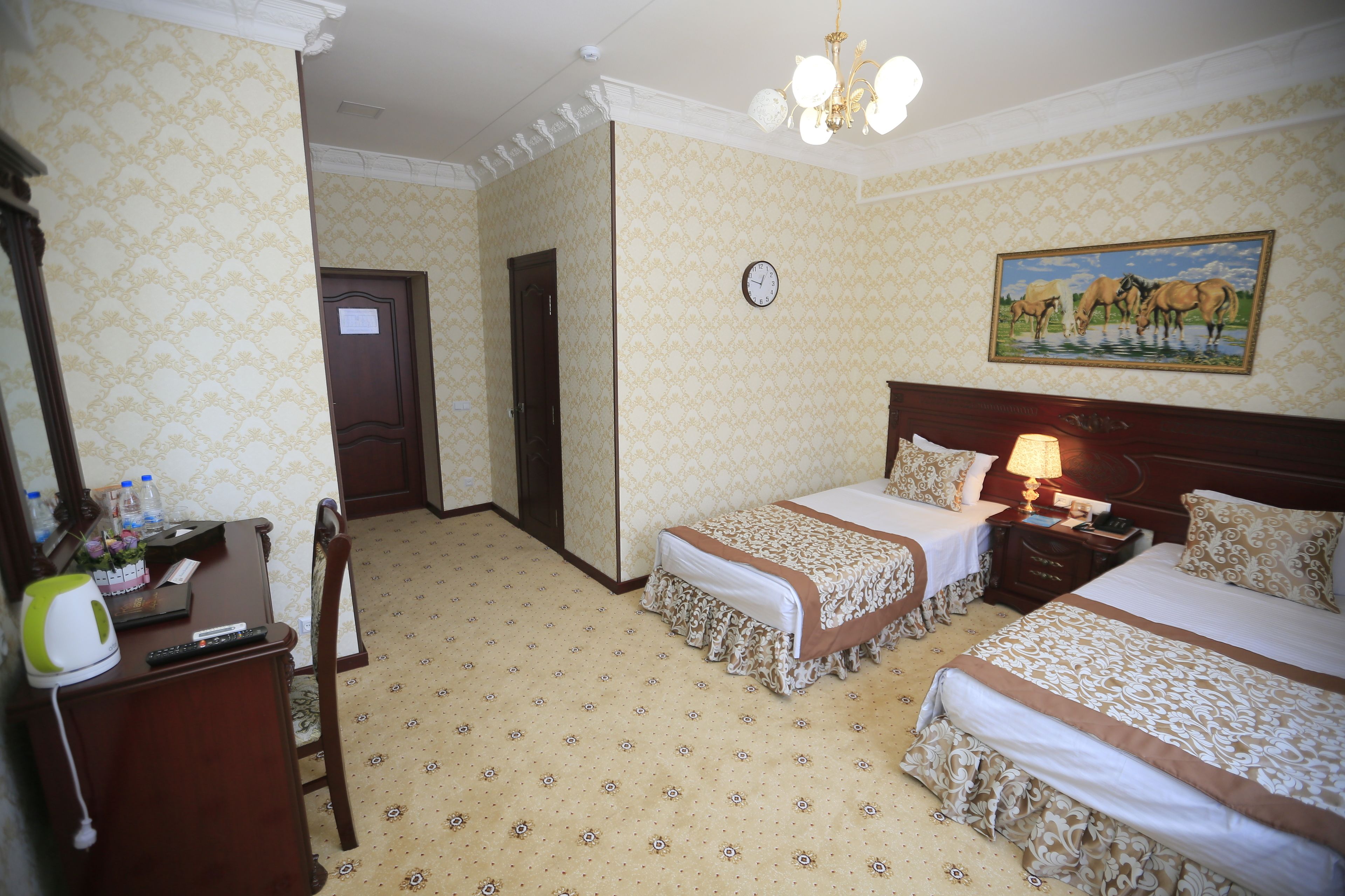 Une chambre d’hôtel avec deux lits, un bureau avec une chaise, une horloge au mur et un tableau au-dessus du lit.